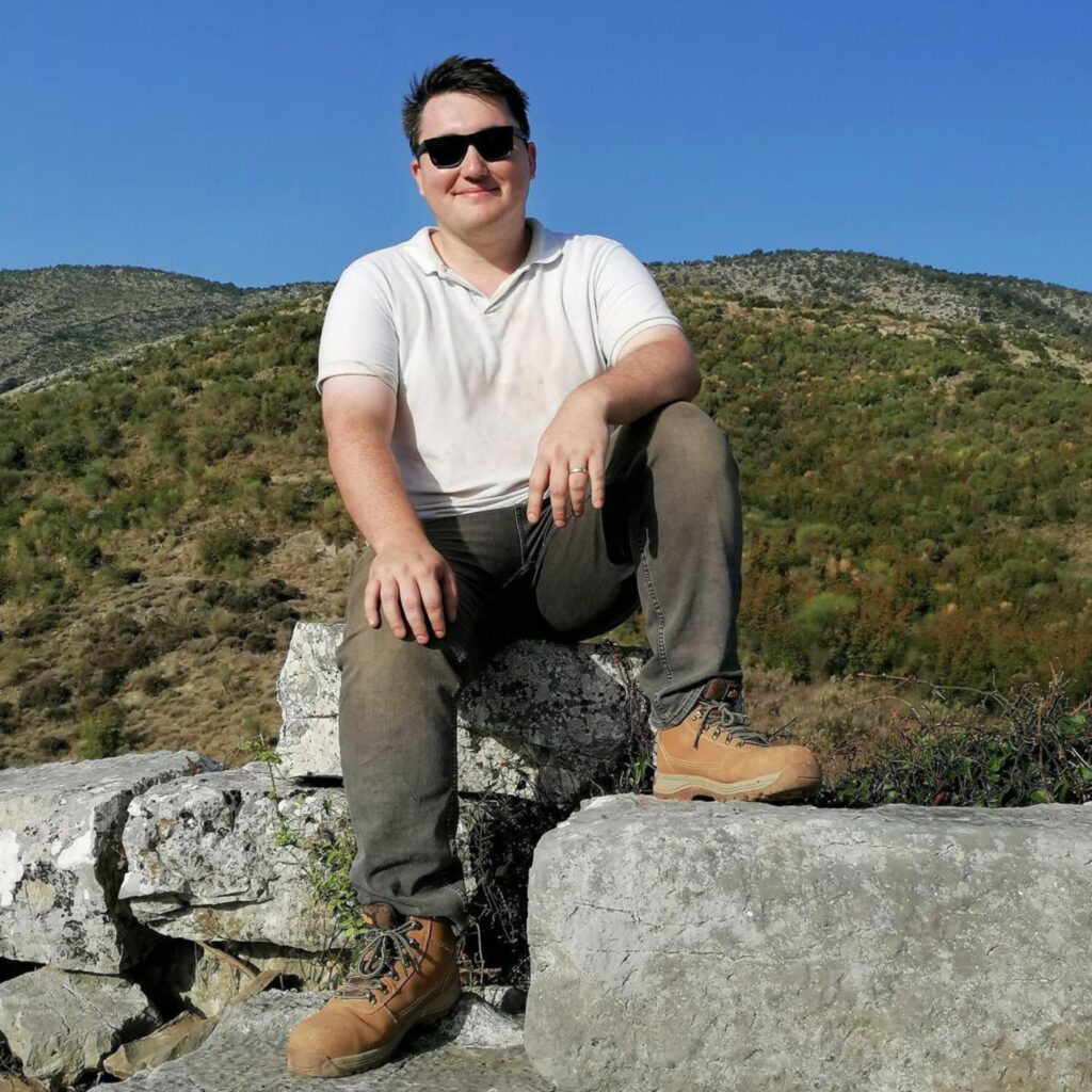 Man wearing white shirt and sunglasses, sitting on top of ancient Greek wall and smiling at camera.