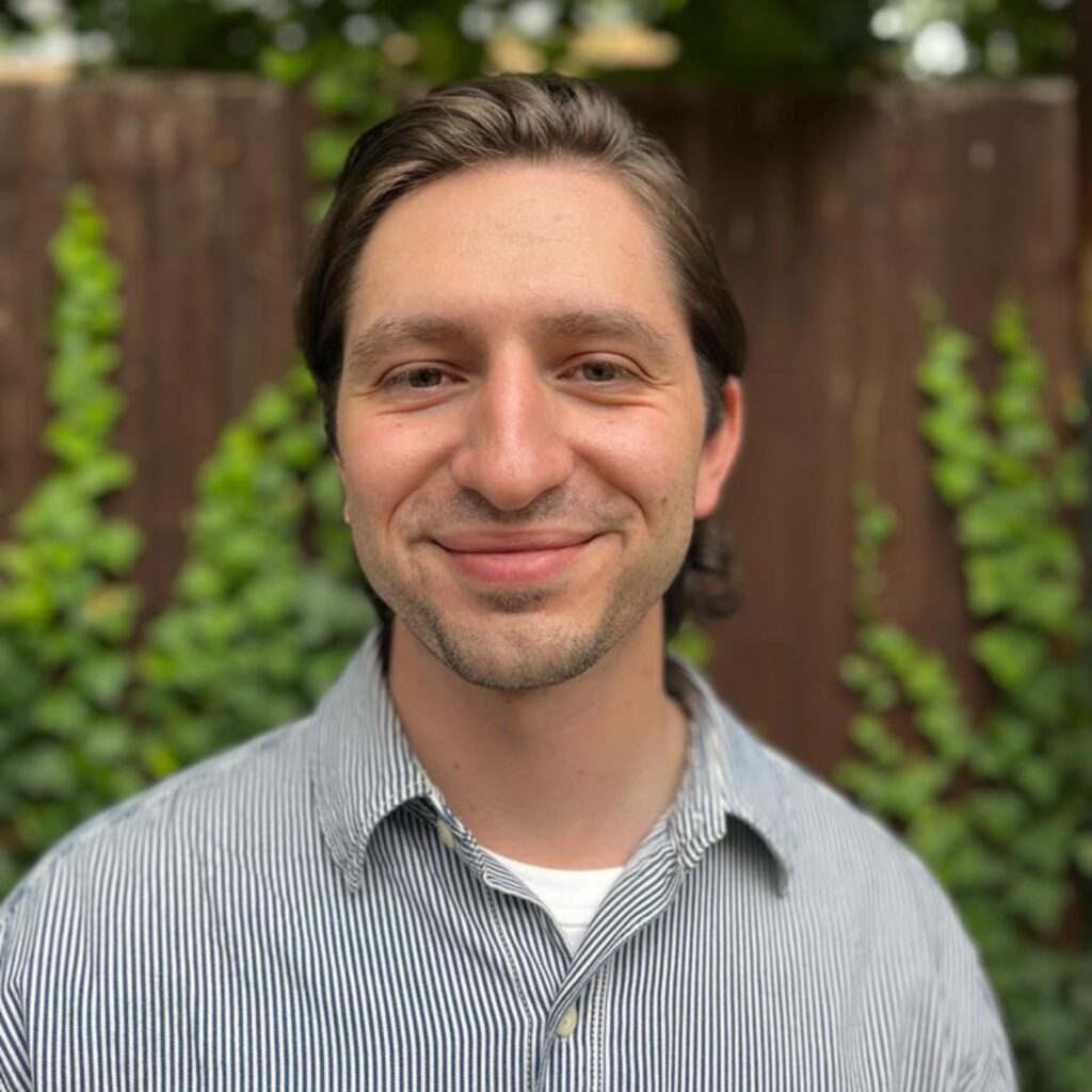 White man standing in front of a fence and shrubbery. He is wearing a striped, blue shirt, and is smiling at camera.