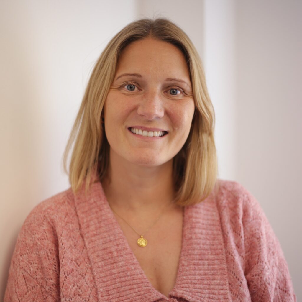 A blonde woman in a pink knitted cardigan smiles at the camera in front of a white wall.