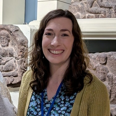 White woman with brown wavy hair smiles at camera. She is standing in front of a museum exhibit of carved Roman stones, and wears a floral dress.